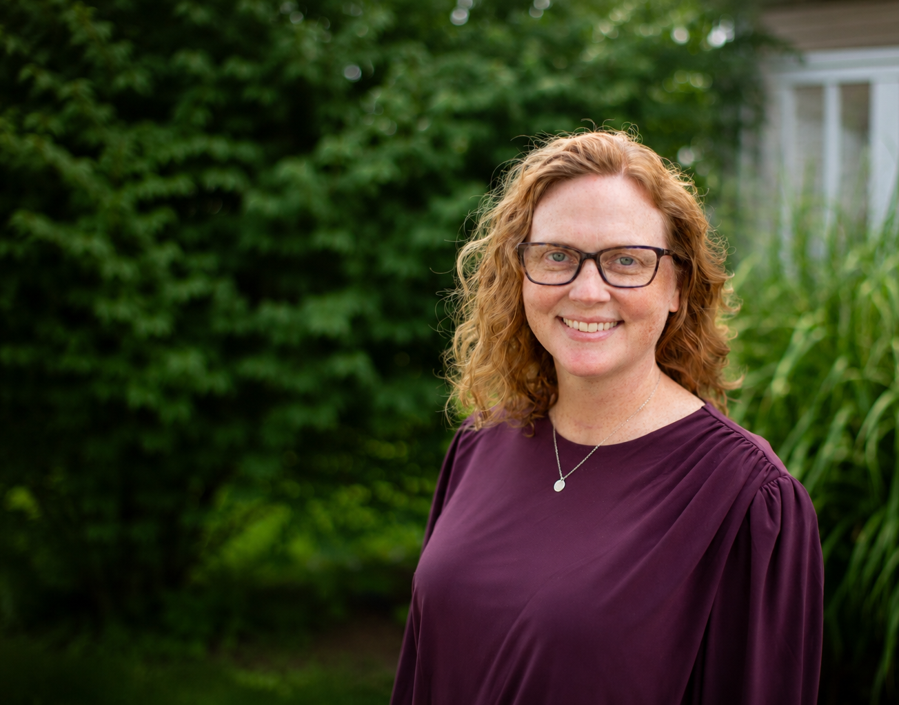Photograph of Kara Reagan, woman with curly red hair and glasses smiling outdoors, wearing a purple blouse and a silver necklace, with green foliage in the background.