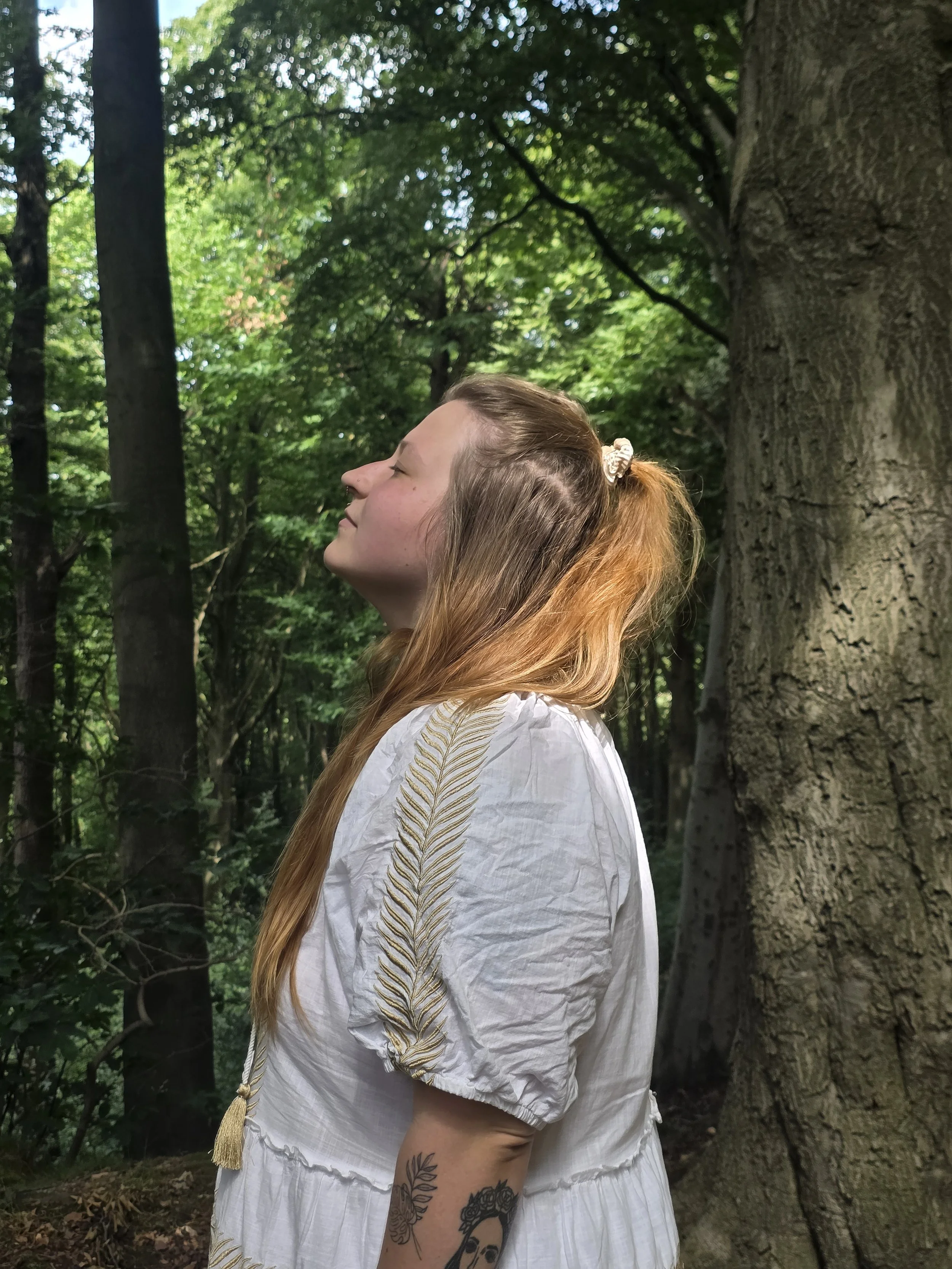 A woman with long red hair, wearing a white garment with gold embroidery, stands outdoors in a forested area with tall trees and green leaves, her face turned to the side with eyes closed.
