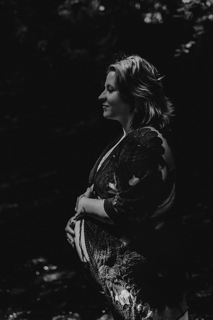 A pregnant woman with shoulder-length hair, standing in water with her eyes closed and hands resting on her belly. She is wearing a lace dress, and the photo is black and white.