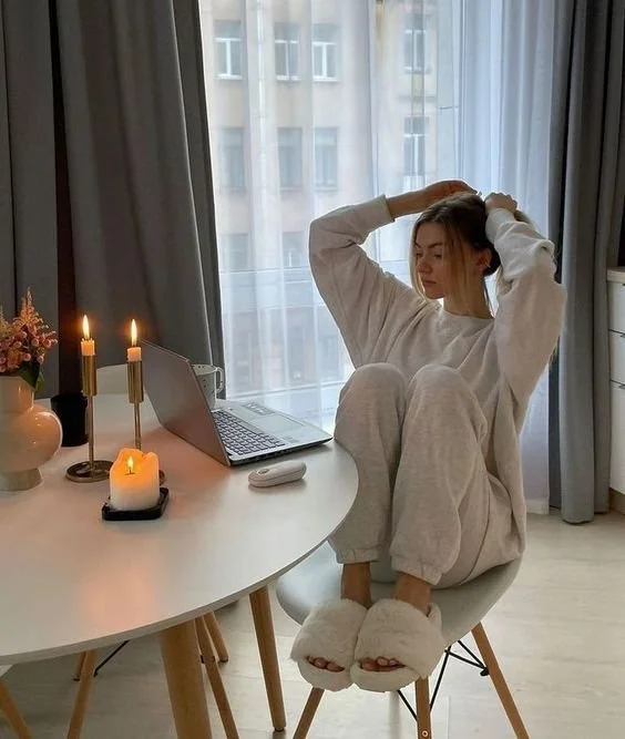 A young woman sitting on a chair at a white table with a laptop, candles, and a flower pot, stretching or adjusting her hair near a window in a cozy room.