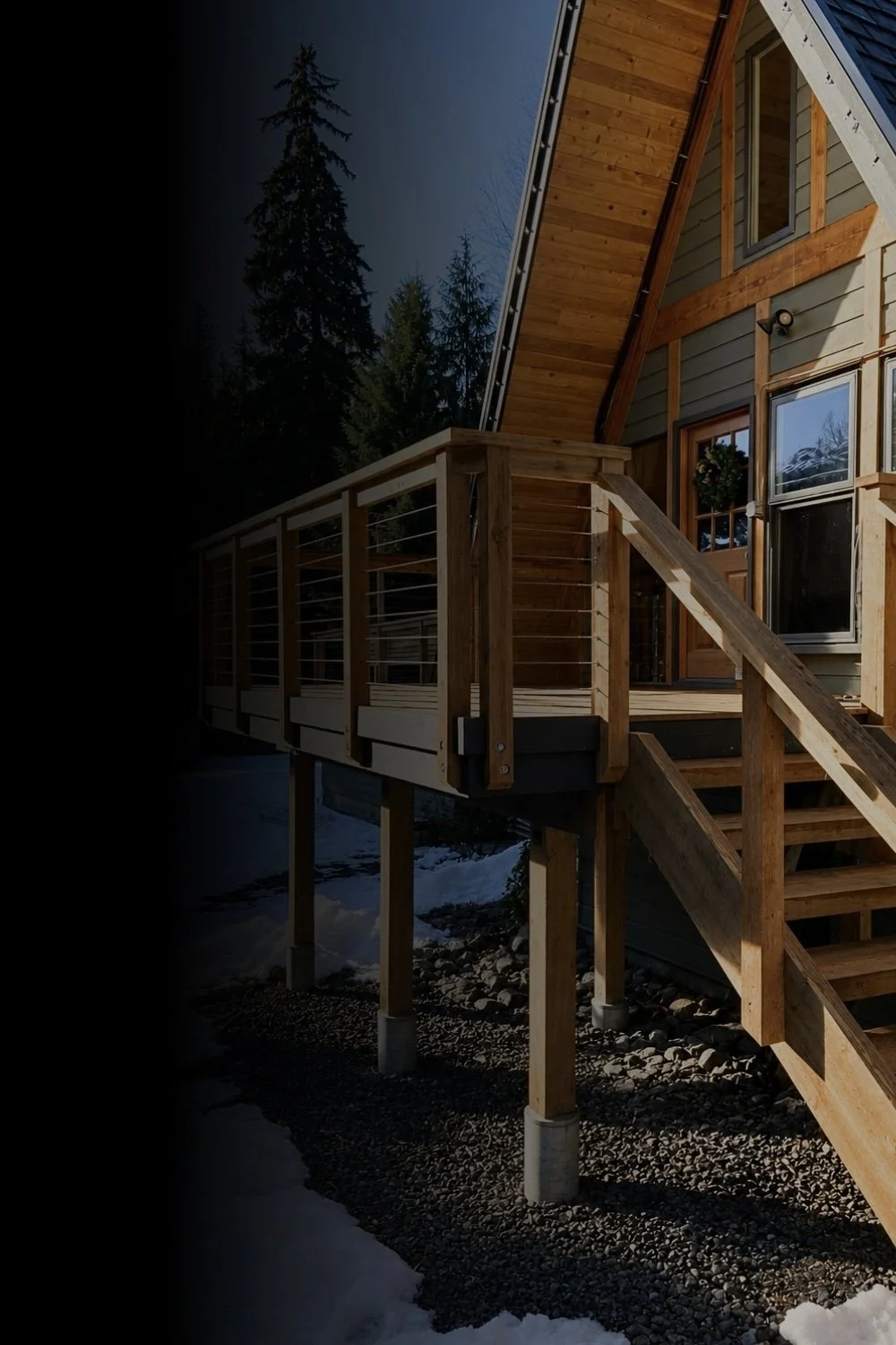 Wooden deck with stairs attached to a house with gray siding and windows, surrounded by tall pine trees and snow on the ground. Mtn Side general contractor bc residential work