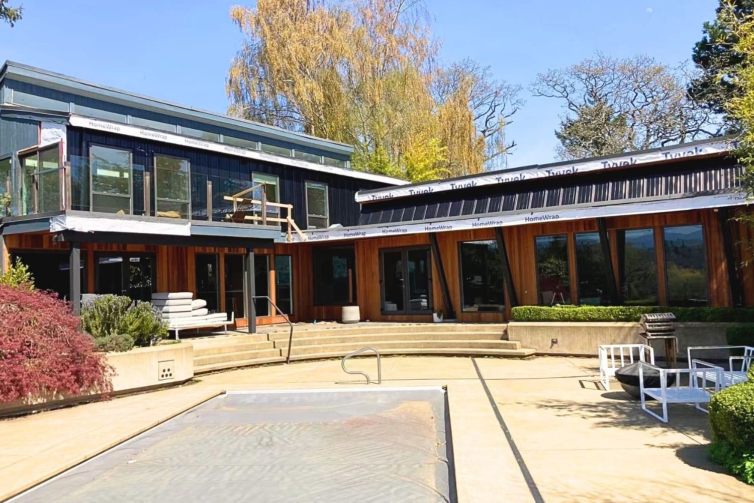 Modern two-story house under construction with large glass windows, wooden exterior, outdoor patio, Construction materials are visible. Mtn Side general contractor bc residential work, cedar siding on home vancouver