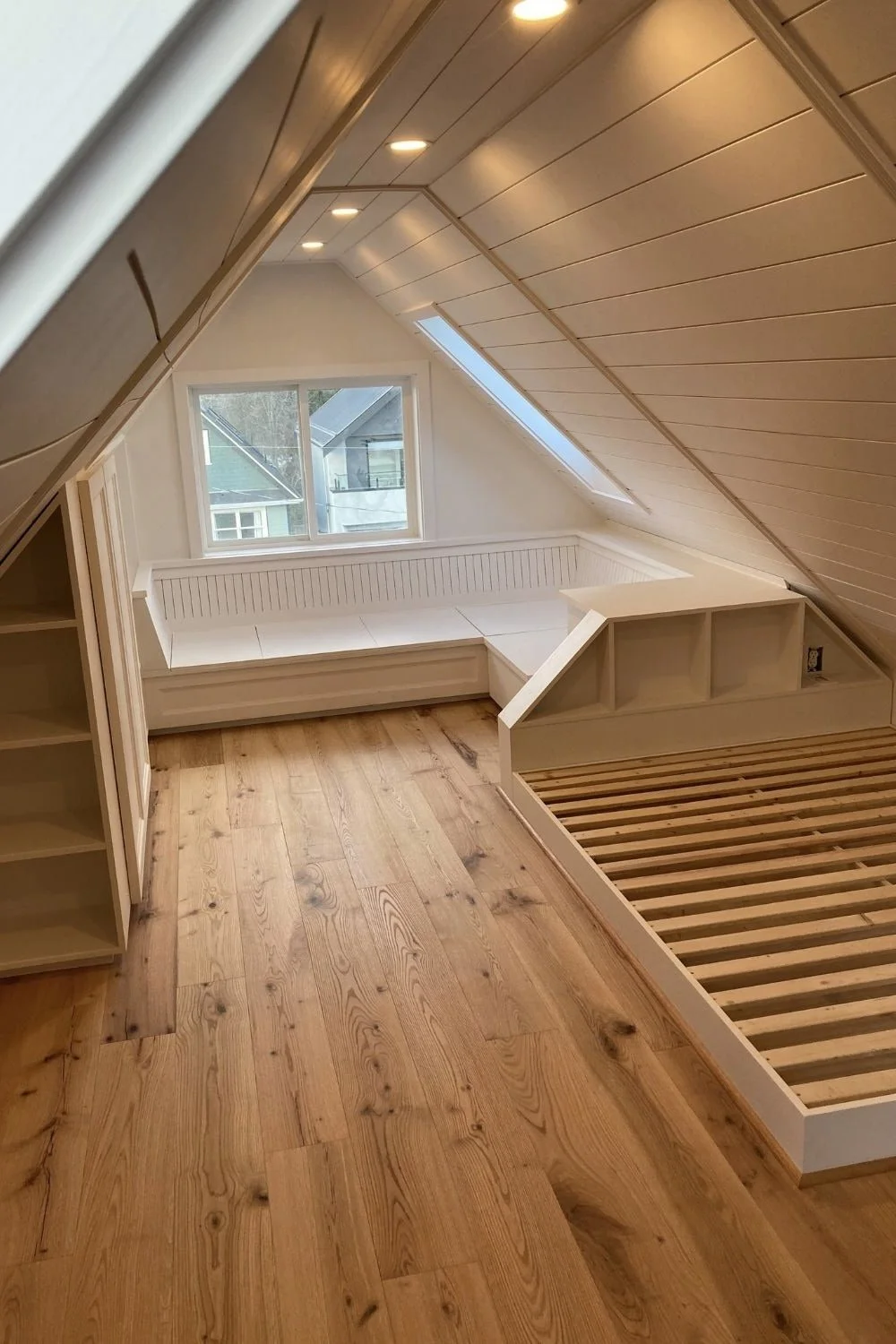 Attic bedroom with a sloped wooden ceiling, a built-in window seat next to a window, and a wooden bed frame without a mattress. Mtn Side general contractor bc residential work