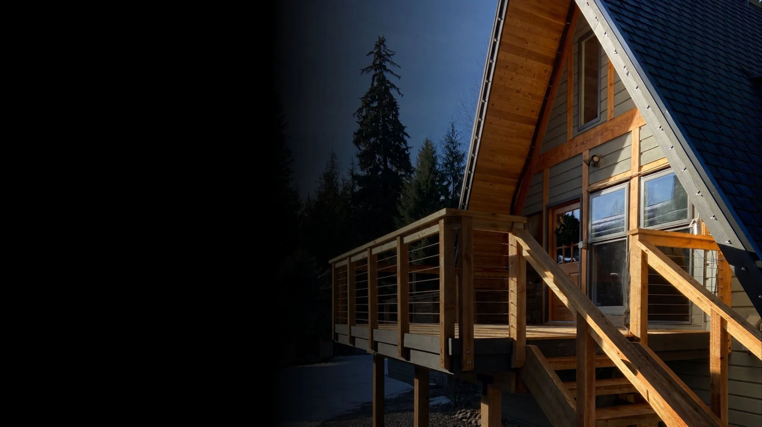 A house with a wooden deck and stairs, surrounded by tall evergreen trees, under a darkening sky.  Mtn Side residential contractor work a-frame deck and railing renovation bc