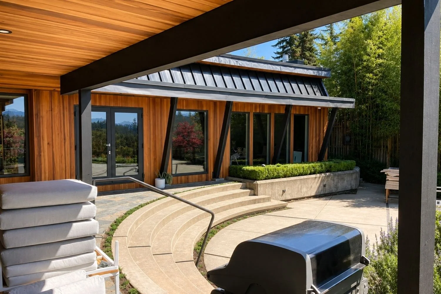 Modern house with wooden exterior, large glass doors, outdoor patio with curved steps, outdoor furniture, a grill, and lush greenery in the background. Mtn Side general contractor bc residential work, cedar siding on home vancouver