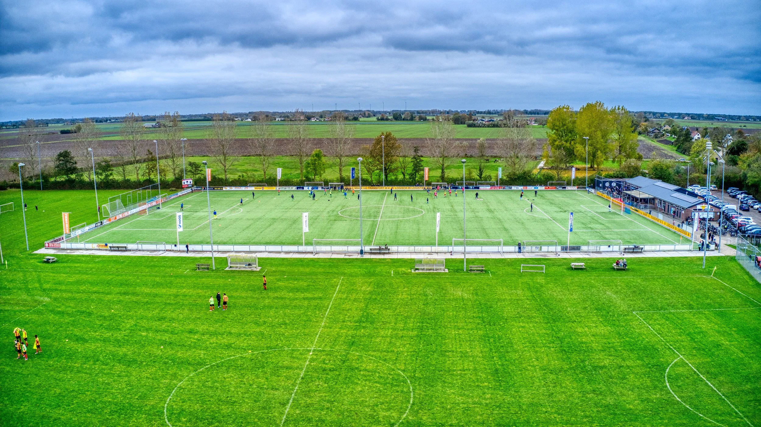 Een voetbalveld met spelers die een wedstrijd spelen, omgeven door een grasveld en landschap met bomen en huizen onder een bewolkte hemel.