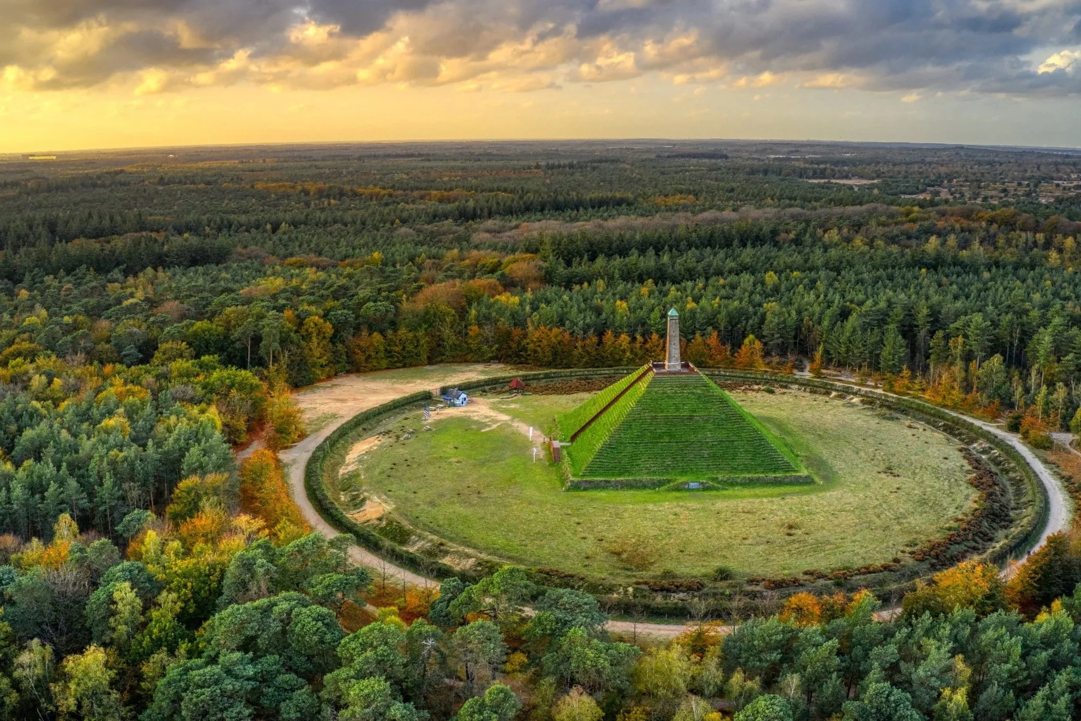 Een uitgestrekt bosgebied met een monumentale structuur in het midden, omringd door paden en groene gebieden, met een uitgestrekt landschap op de achtergrond onder een bewolkte hemel.
