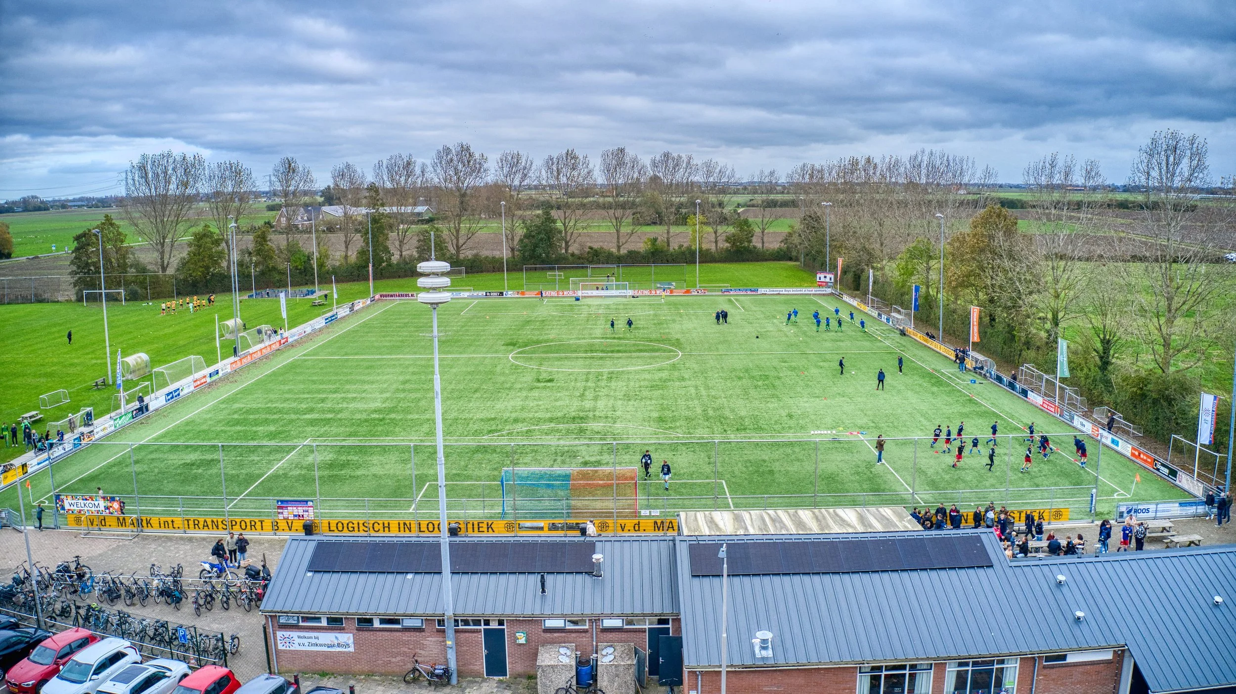 Een voetbalveld met spelers die trainen of opwarmen, omgeven door een hek en bomen in de achtergrond, onder een bewolkte lucht.