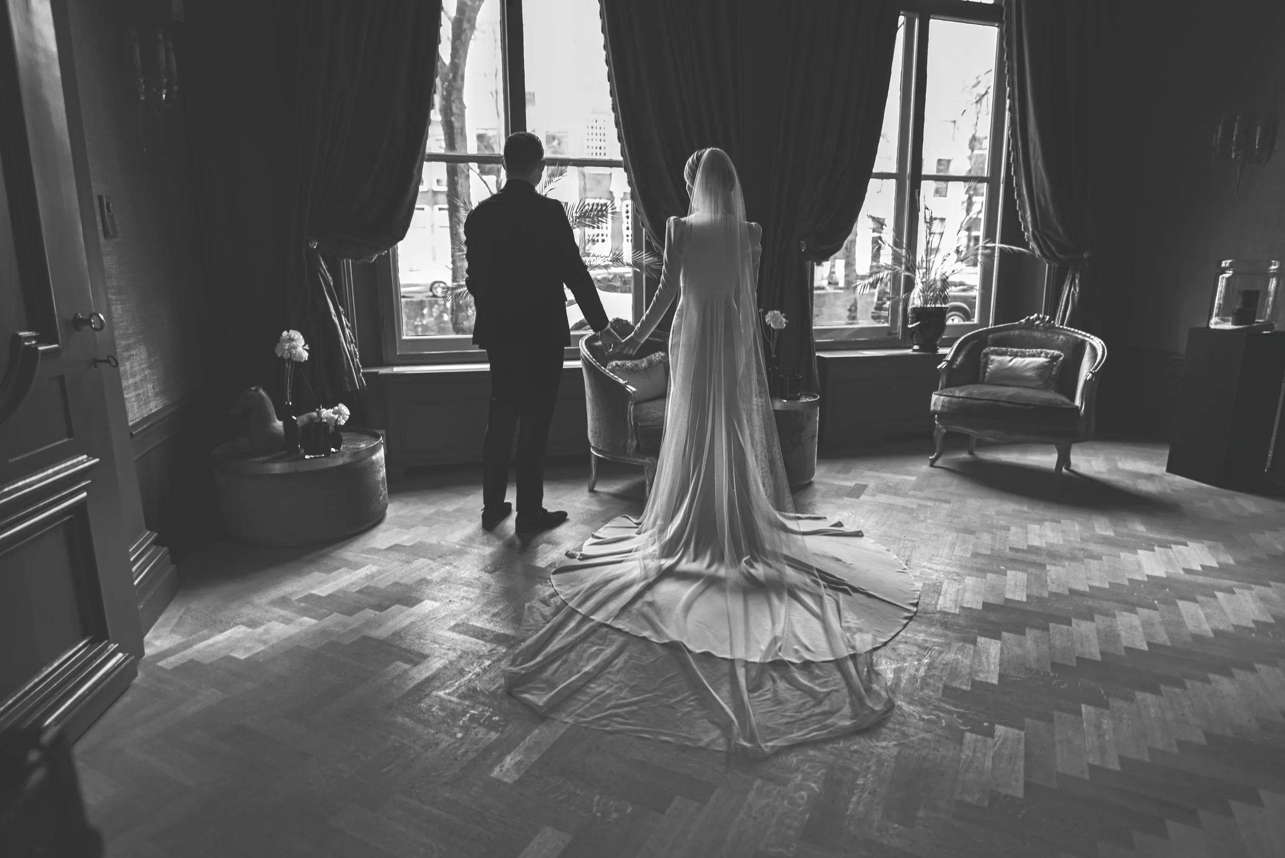 Een zwart-wit foto van een bruid en bruidegom die hand in hand voor een groot raam staan, met gordijnen aan beide zijden, in een elegante binnenruimte.