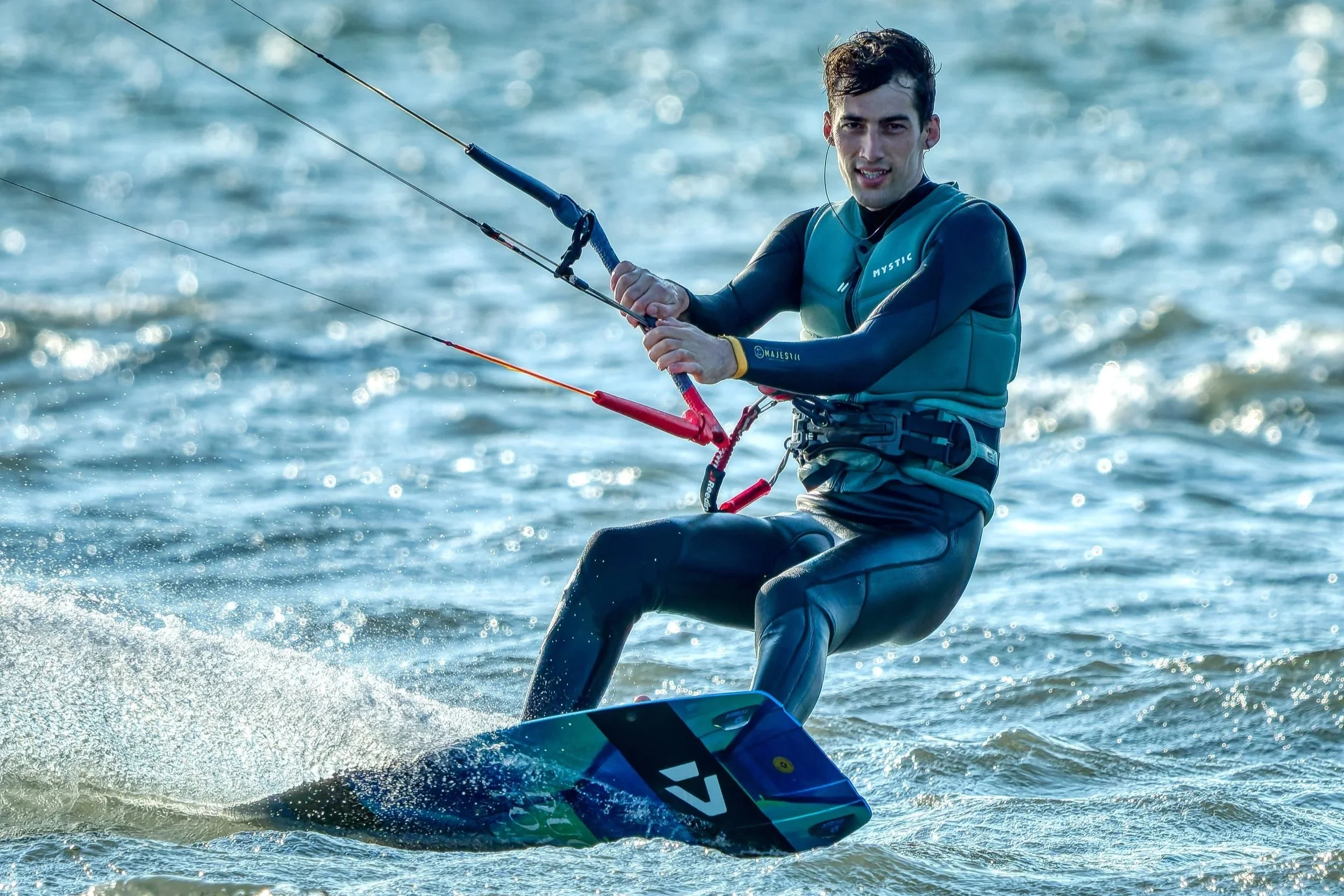 Man die windsurft op de zee, met een blauwe windsurfboard en een wetsuit, terwijl hij zich vasthoudt aan de mast met het zeil.