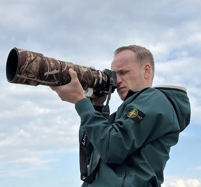 Man met een groen jack gebruikt een telelens om door te kijken, tegen een bewolkte lucht