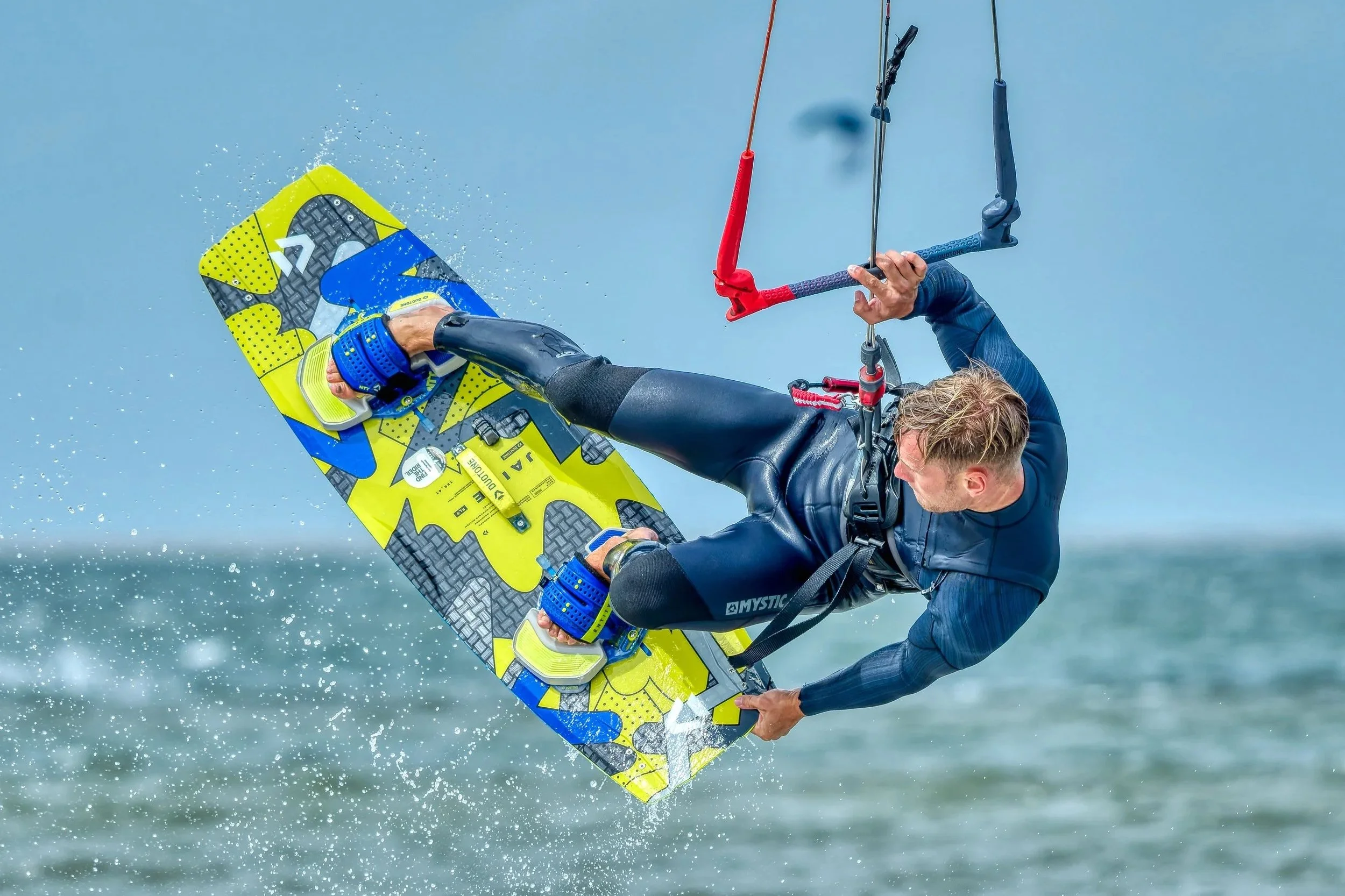 Een surfer die een sprong maakt uit het water met een geel en blauw surfboard, terwijl hij een wetsuit draagt.