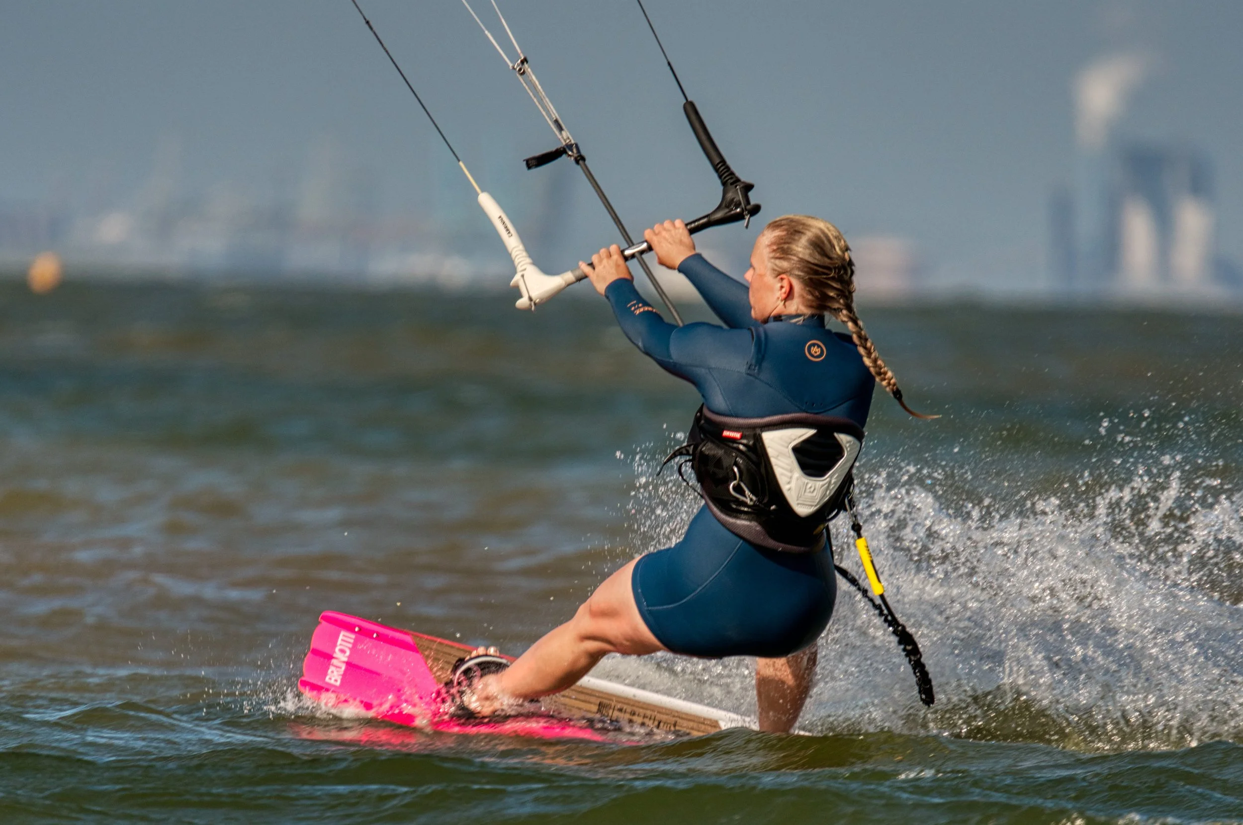 Een vrouw die windsurft op de zee, gekleed in een blauwe wetsuit en met lang blond haar in een vlecht.