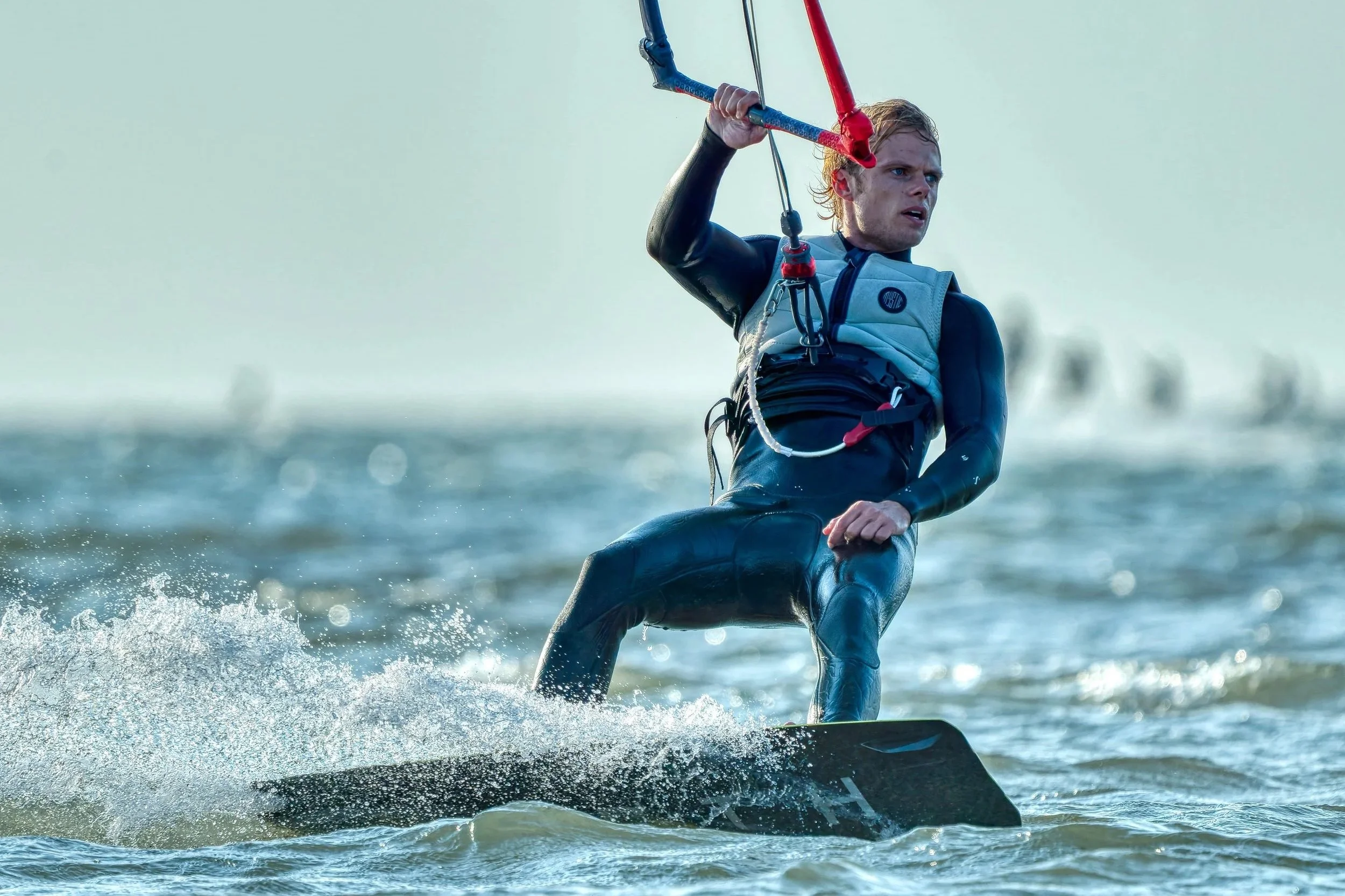 Man die kitesurft op zee, gedragen in een wetsuit, met een blauwe en rode controlenbar in zijn handen, water spettert rond hem.