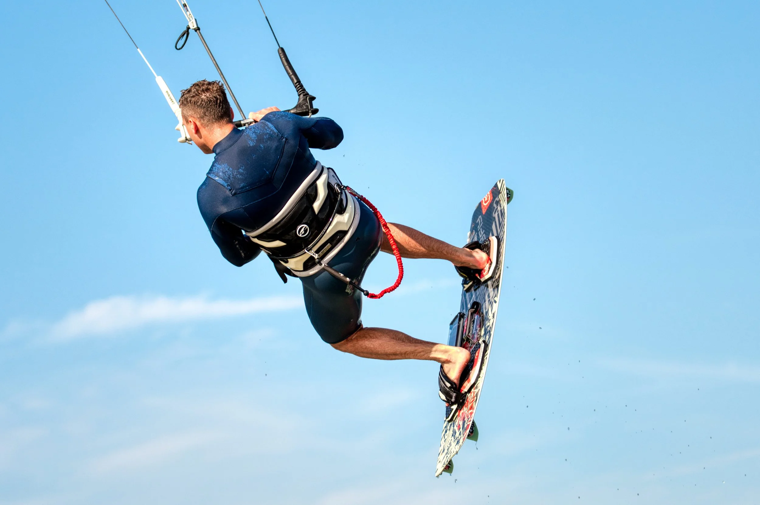 Man die kitesurft in de luchtruimte, gekleed in een wetsuit en met een kite control bar, onder een heldere blauwe hemel.