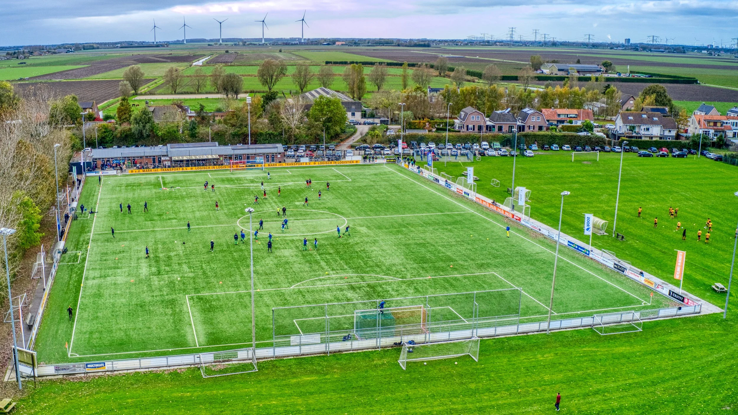 Een voetbalveld met spelers die trainen en oefenen, omgeven door een groene omgeving met huizen en weilanden in de achtergrond, onder een bewolkte hemel.