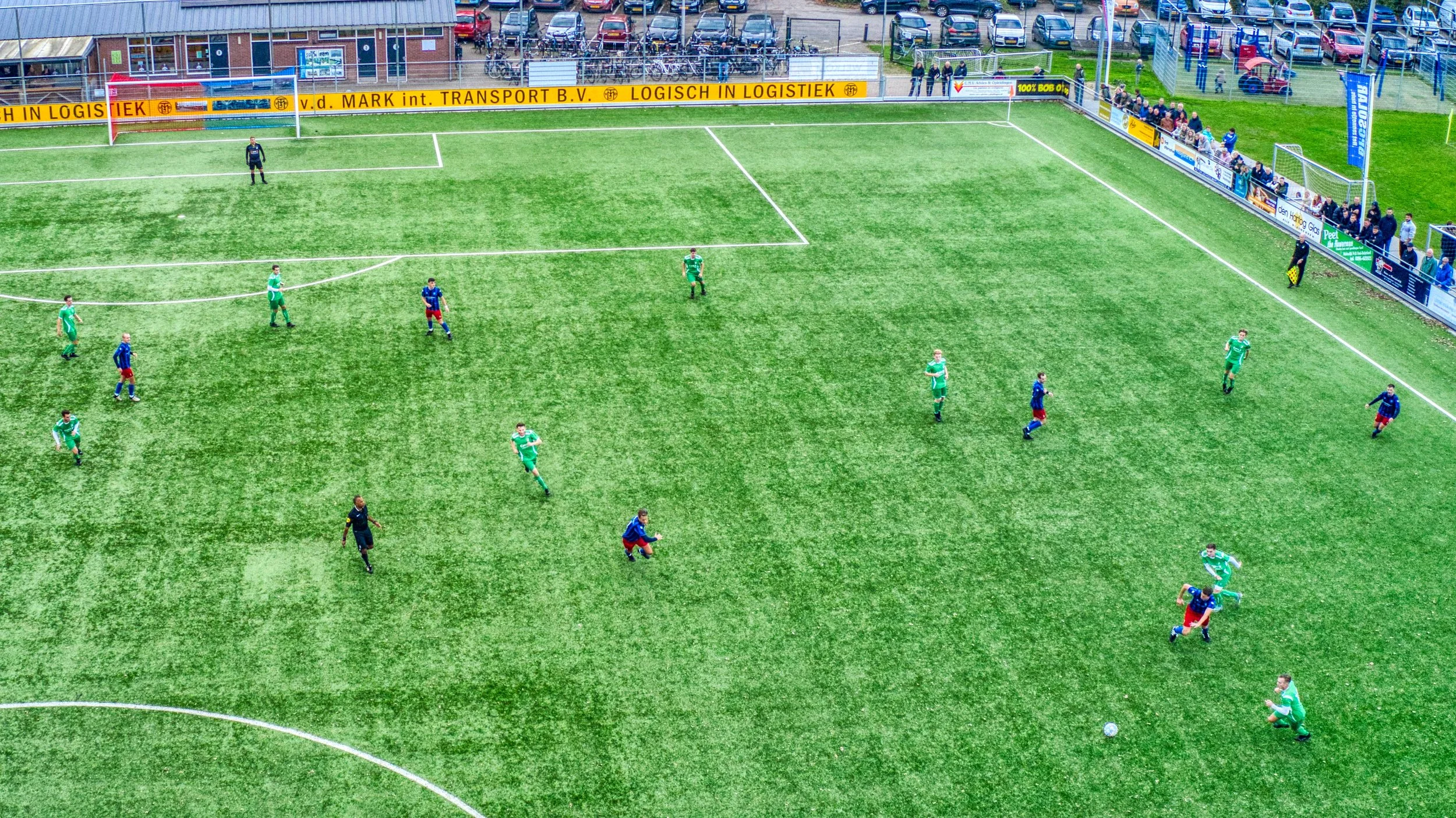 Een voetbalwedstrijd is bezig op een kunstgrasveld, met spelers in groen en blauw tenue en een keeper in zwart, terwijl een kleine groep toeschouwers langs de kant kijkt.