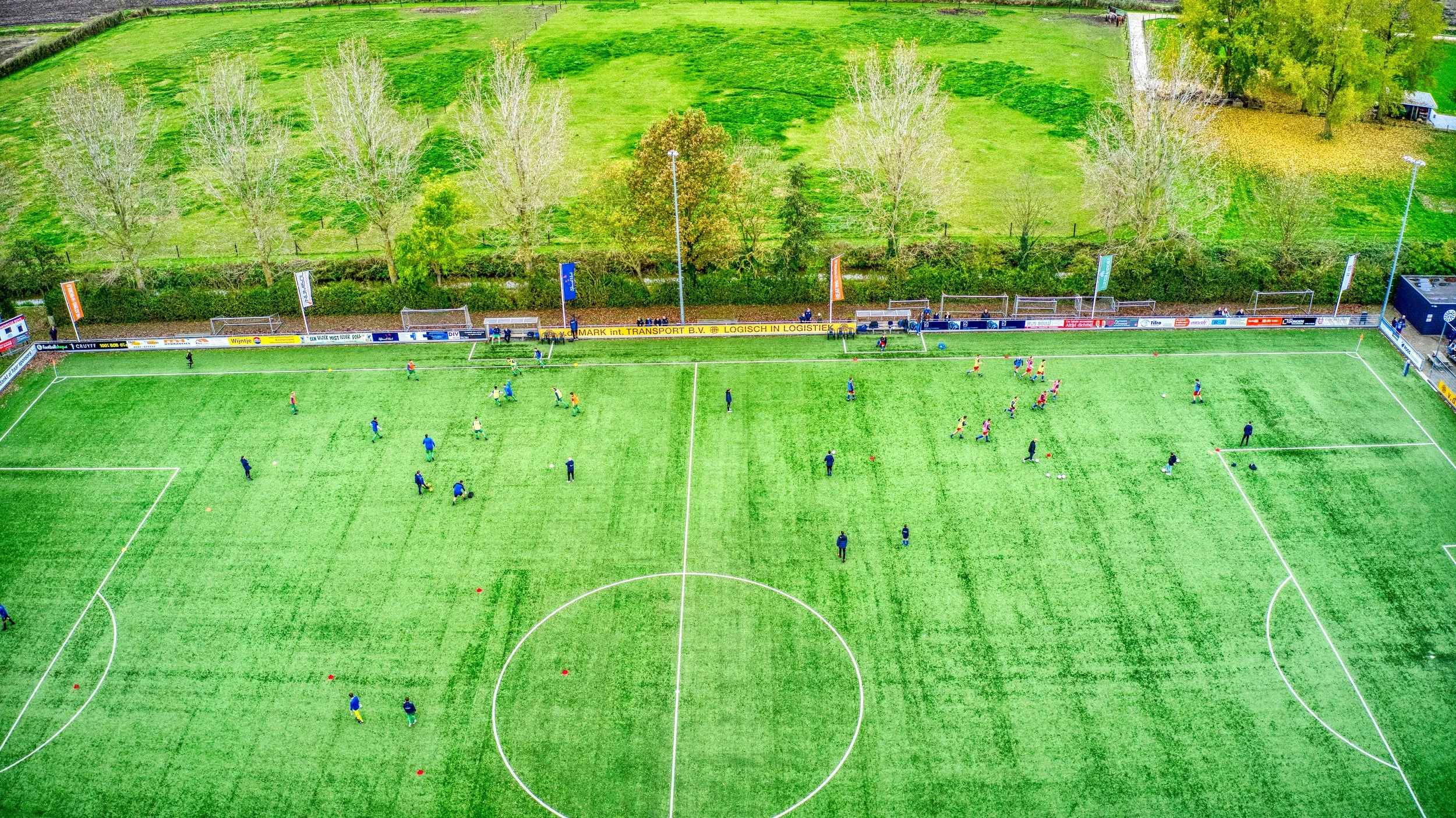 Luchtfoto van een voetbalveld met spelers die oefenen, omringd door een groene grasweide en bomen.