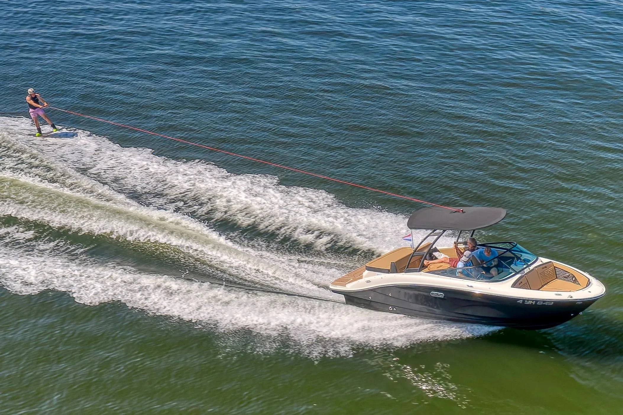 Persoon wakeboarden achter een speedboot op het water, met blauwe water en een groep mensen in de boot.