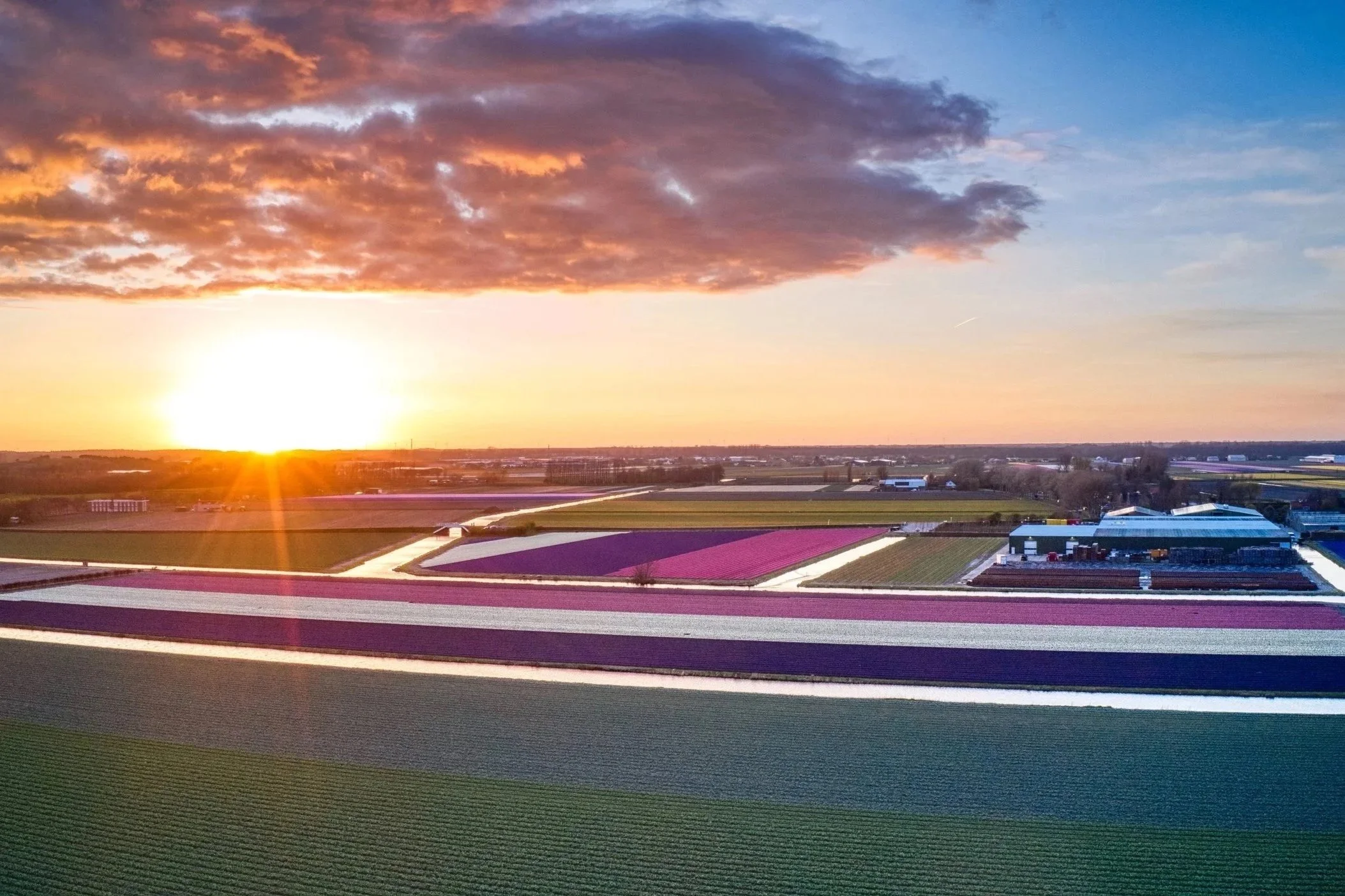 Uitzicht over een landschap met velden en gebouwen bij zonsondergang, met een kleurrijke lucht en bewolking.
