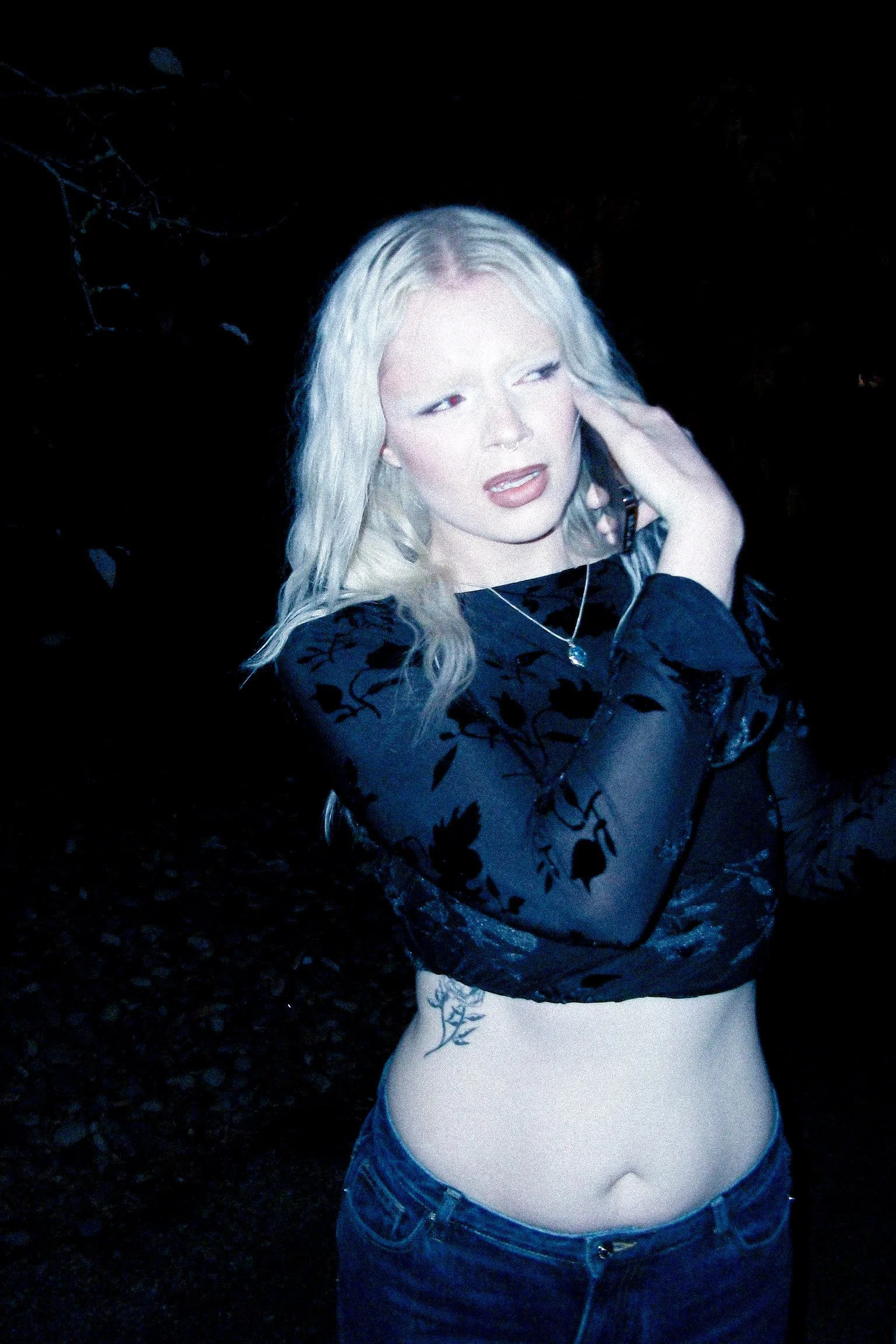 A woman with long blonde hair wearing a black floral top and blue jeans, standing outdoors at night.