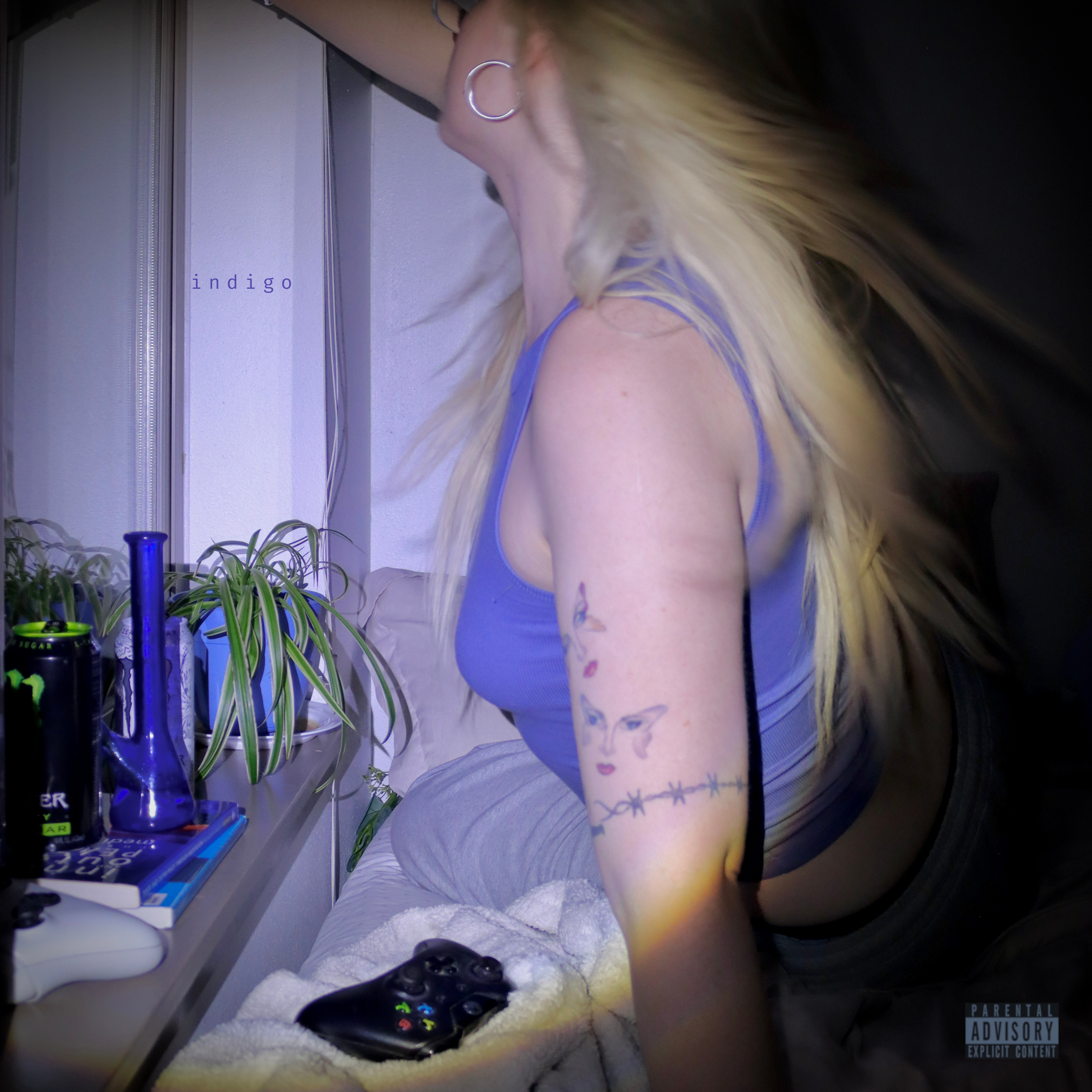 A person with long blonde hair, wearing a blue tank top, sits at a desk with various gaming controllers and a PlayStation console in front, behind some potted plants and shelves with bottles and other items, in a room with a window.