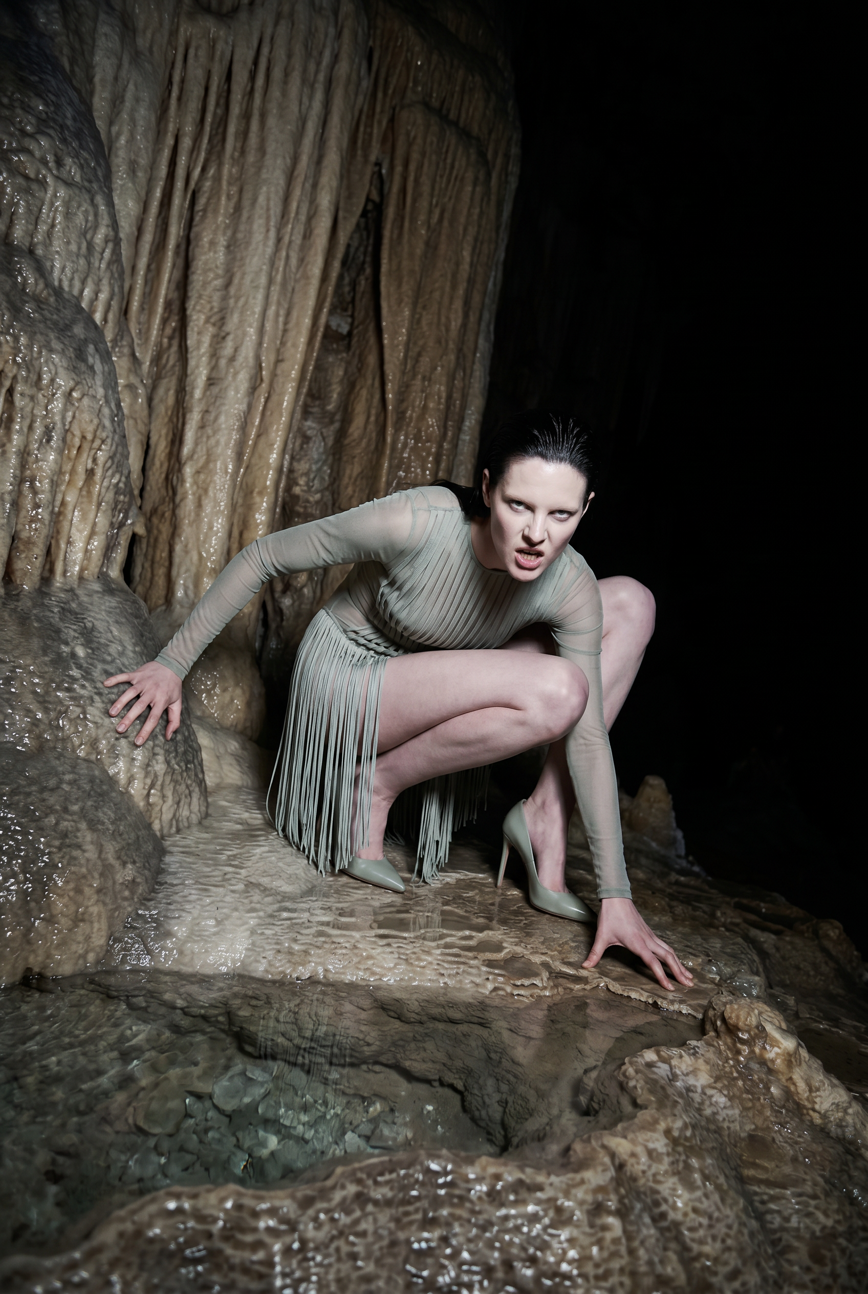 A woman with dark wet hair, wearing a beige dress with fringes and high heels, crouching on a wet, rocky surface in a cave with stalactites and stalagmites.