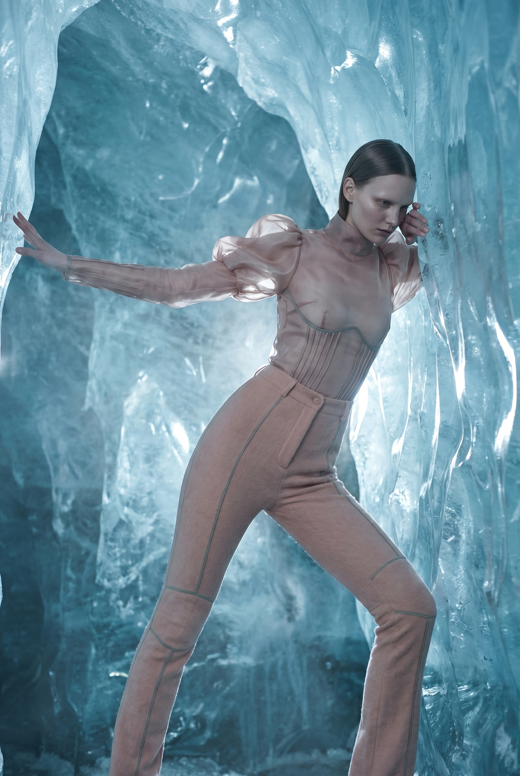A woman wearing beige pants and a shiny beige top poses inside an ice cave, with her right hand pressed against the ice wall and her left hand near her face, looking intense.