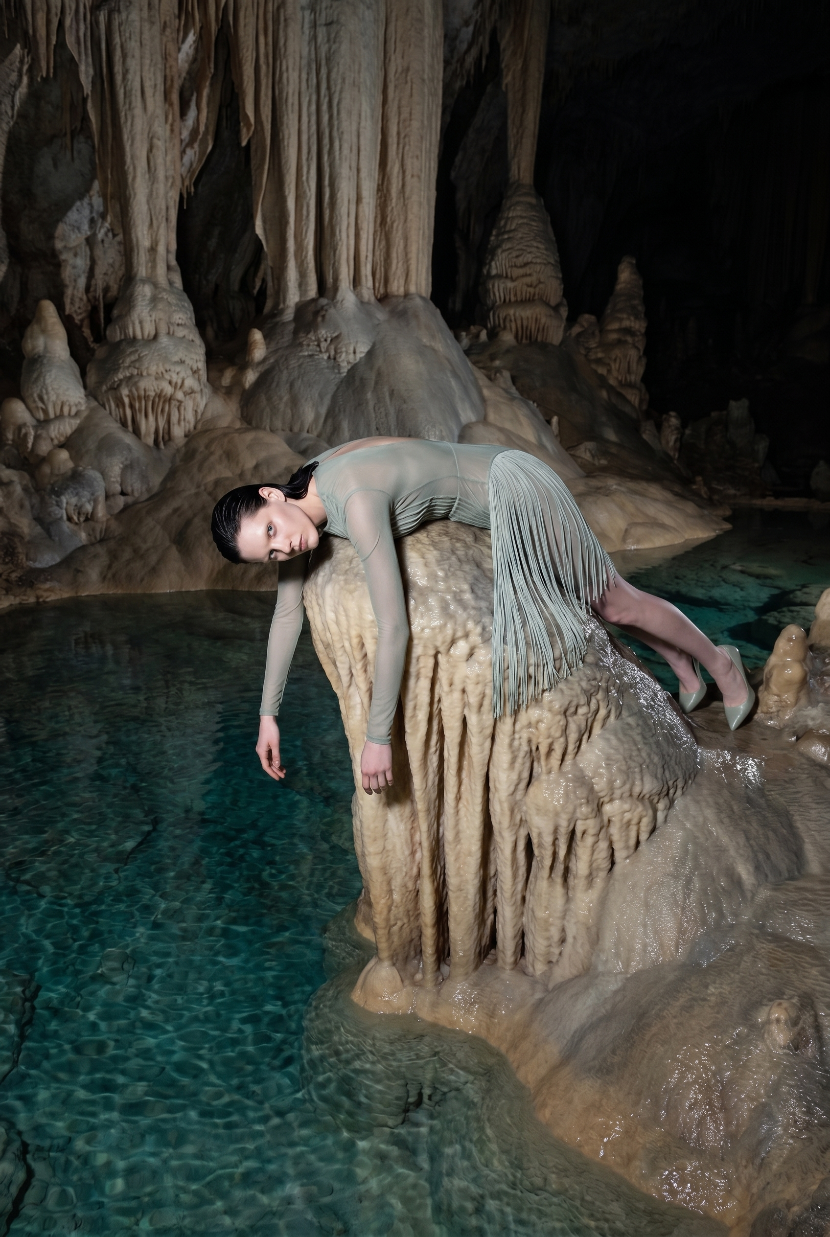 A woman in a sheer, long-sleeved dress with fringe is posing on a large, stalagmite formation inside a cave with hanging stalactites and a pool of water at the base.
