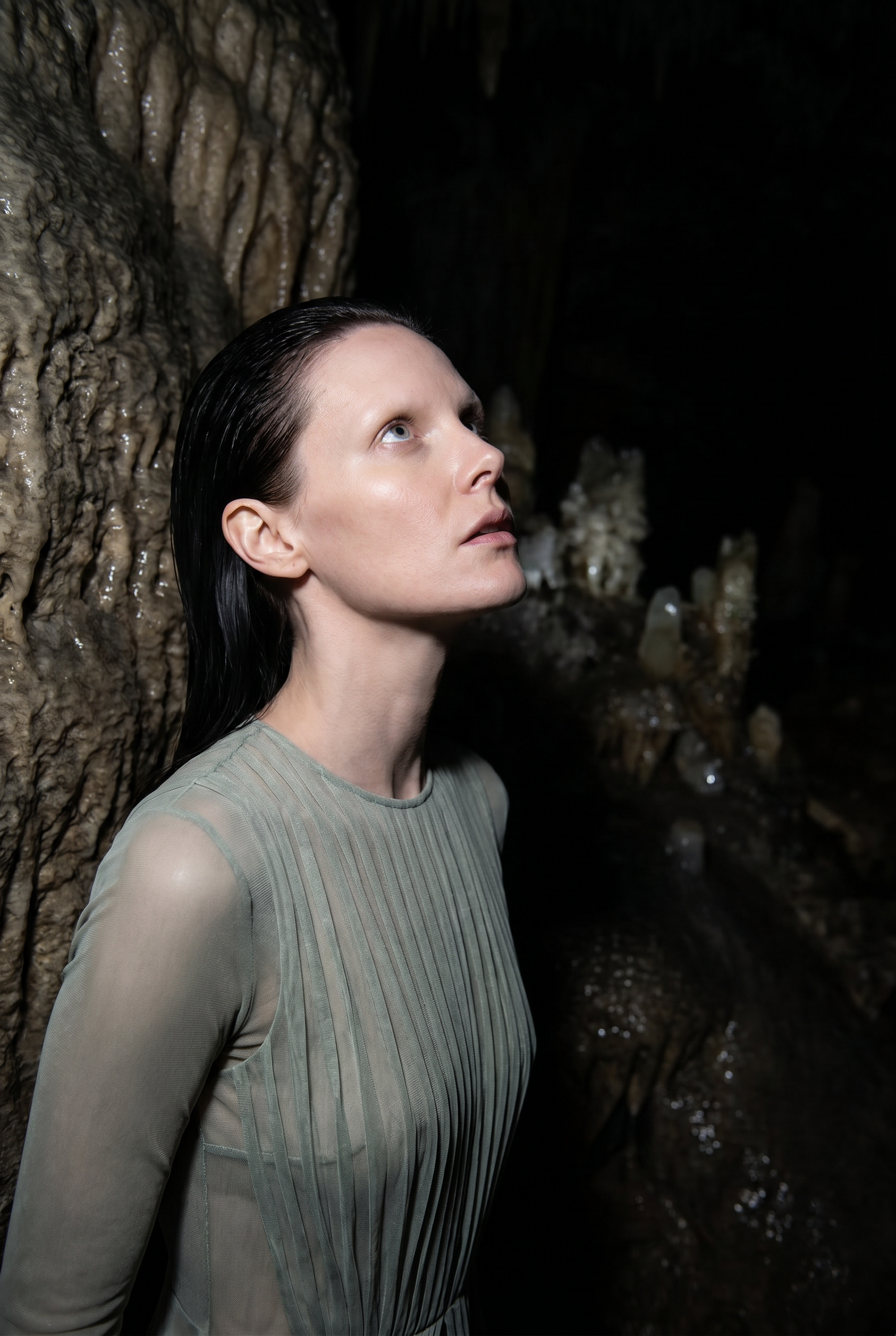 A woman with dark, wet hair looking upward in a dark, cave-like setting with textured rock formations behind her.