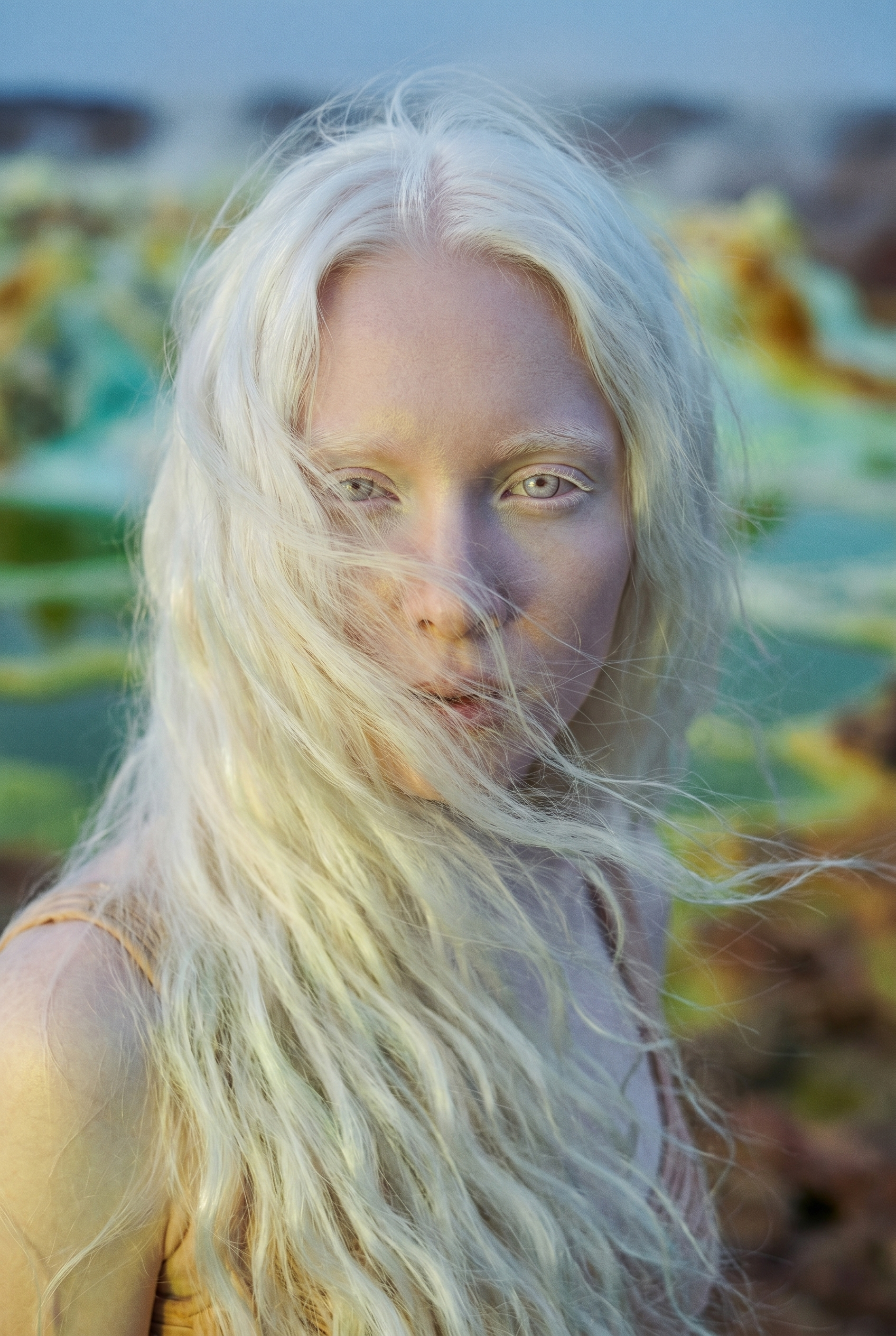 Close-up of a woman with long, platinum blonde hair, light-colored eyes, and a natural expression, standing outdoors near a body of water and foliage.