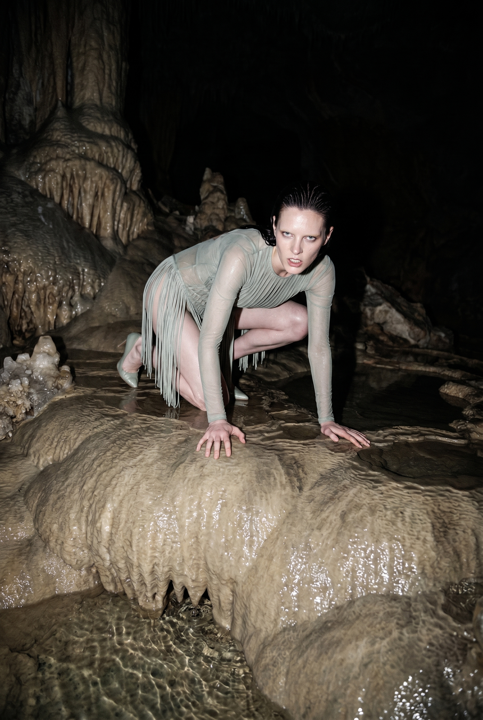 A woman with wet hair and pale green dress crouches on a rocky surface inside a dark cave, surrounded by stalactites and stalagmites.