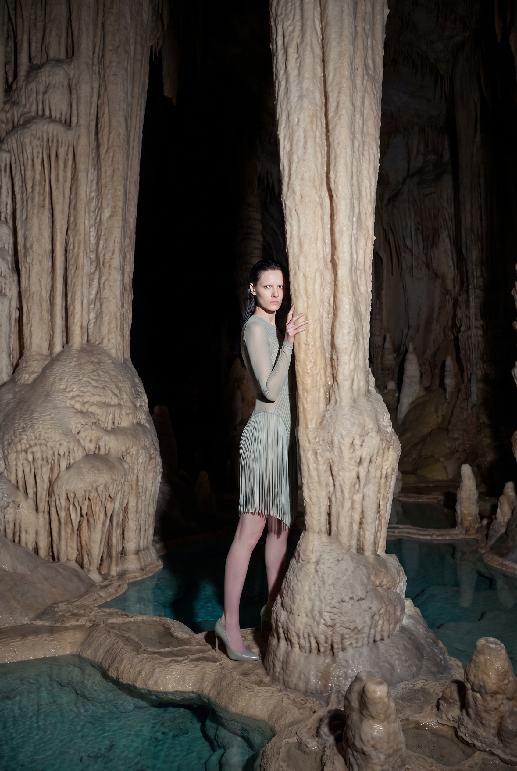 A woman in a light green dress and high heels standing among beige stalactites and stalagmites inside a cave with a pool of water at the bottom.