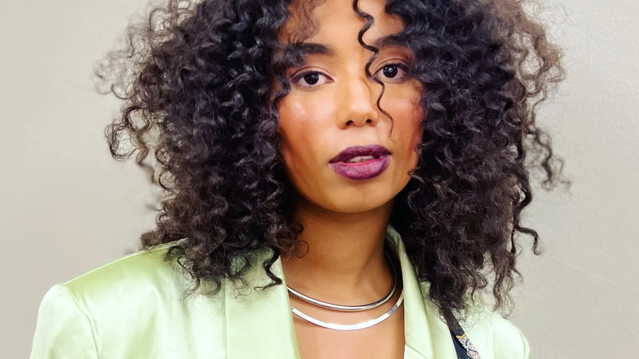 A woman with dark, curly hair and light makeup, wearing a green jacket, silver necklace, and purple lipstick, standing in front of a plain light-colored wall.
