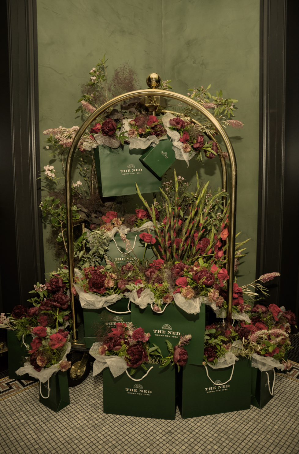 Display of floral arrangements and shopping bags from The Ned hotel in New York, arranged with various flowers and plants against a green wall.
