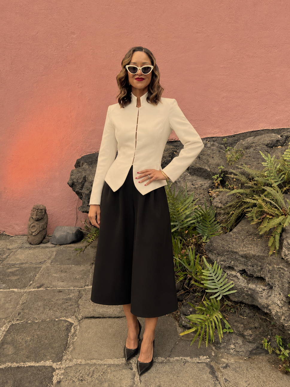 A woman dressed in a white blazer with black wide-leg pants, black high heels, and white cat-eye sunglasses stands against a pink wall with rocks and plants behind her.