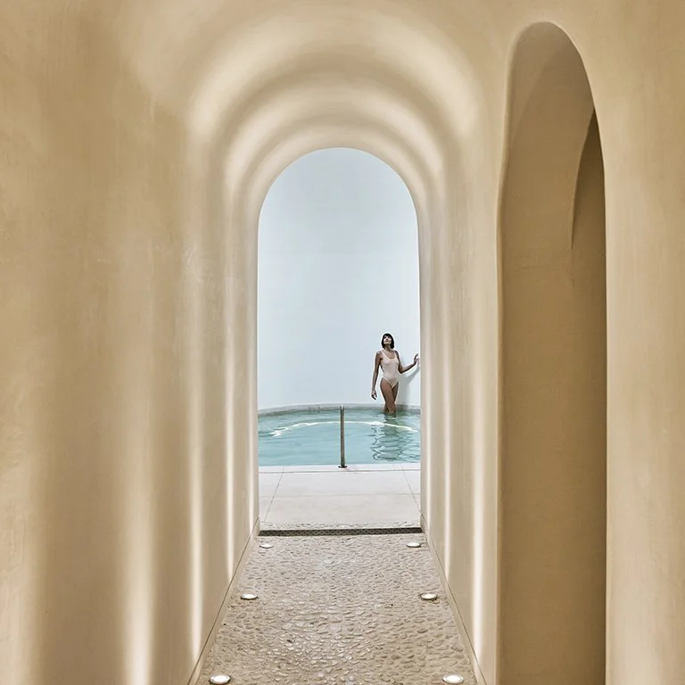 A woman in a swimsuit standing in an indoor swimming pool, viewed through an arched hallway of a spa.