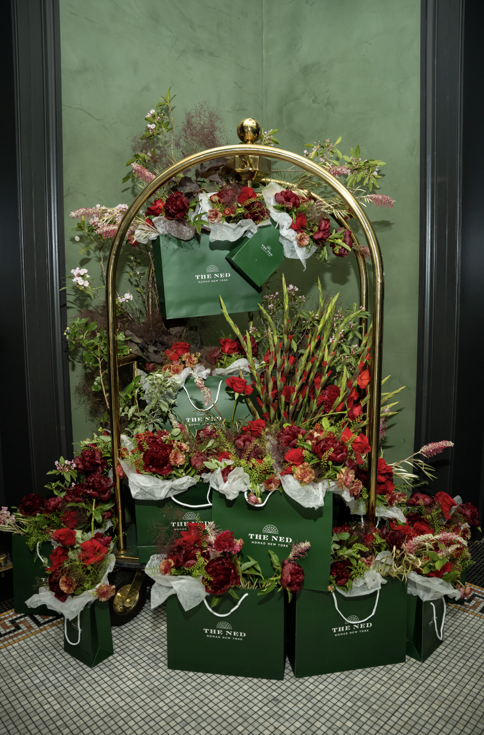 A display of red and pink flowers arranged on a gold luggage cart, with green shopping bags from The Ned hotel, set against a green wall.