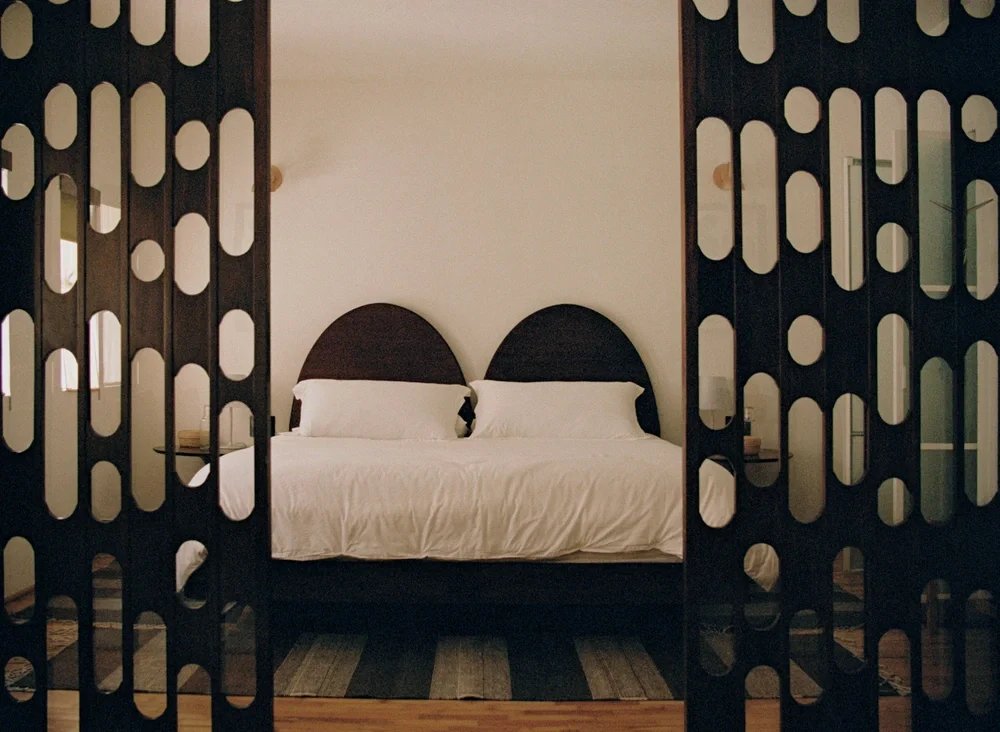 A bedroom with a bed featuring two dark semi-circular headboards, white bedding, and two pillows, viewed through decorative black wooden room dividers.