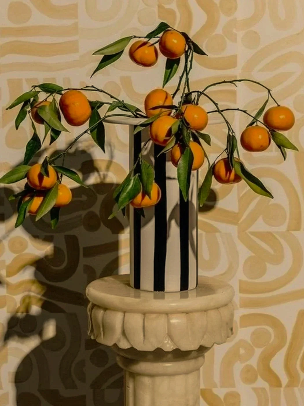 Decorative arrangement of artificial oranges with green leaves in a black and white striped vase on a beige pedestal with a patterned background.