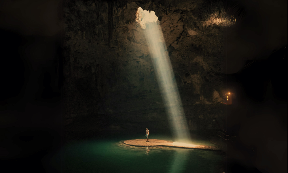 A person stands on a small sandy island inside a dark cave with a tall opening from which a beam of sunlight shines through, illuminating the island and the person.