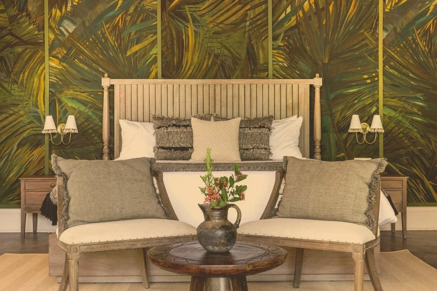 Bedroom with a wooden bed frame and a tropical leaf patterned wallpaper. Two beige nightstands with white lamps are on either side of the bed. A beige sofa with large pillows is in front of the bed, and a vase with flowers is on a round wooden table in front of the sofa.