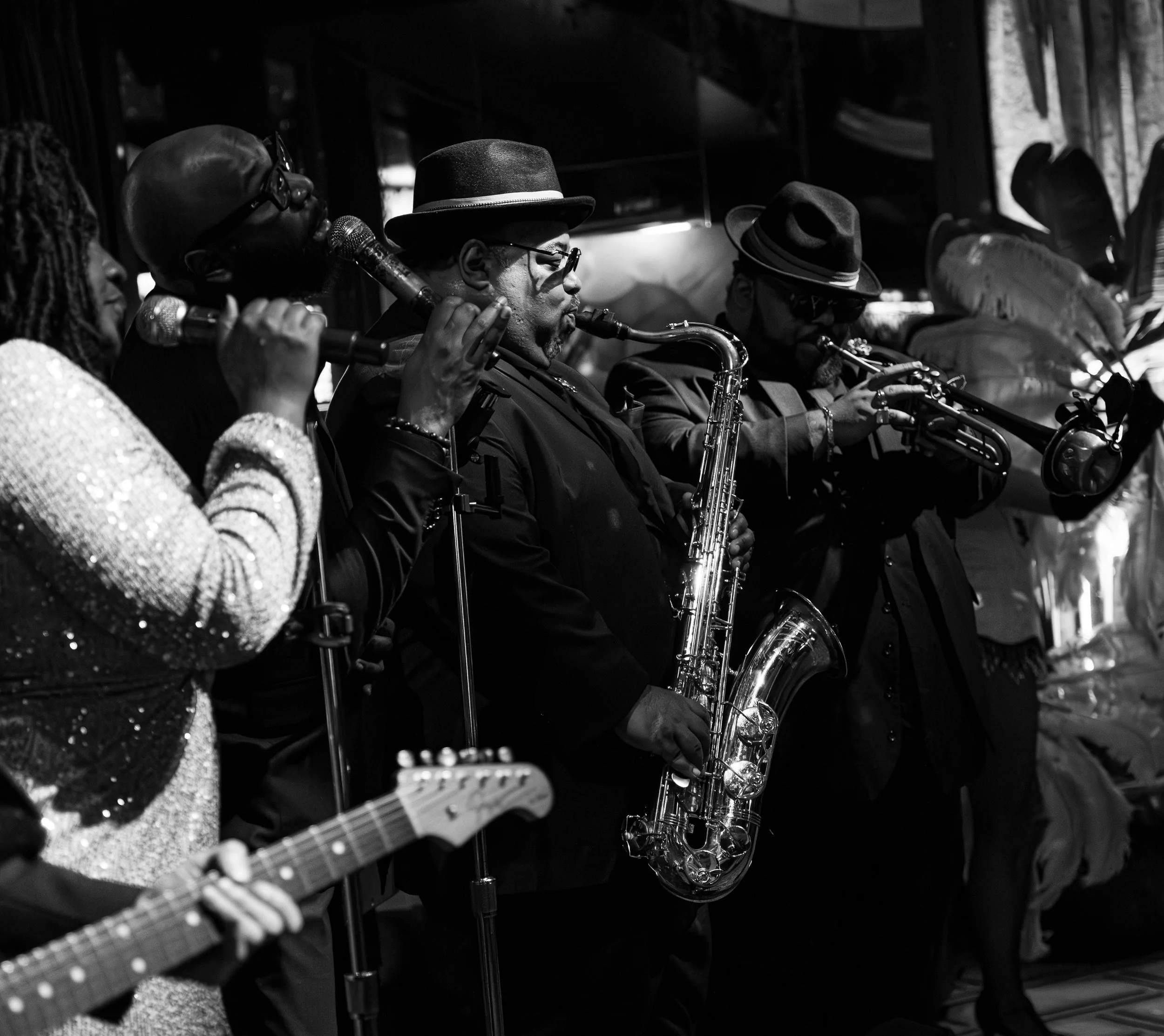 A black and white photo of a jazz band performing, featuring musicians playing saxophones, a guitar, and an electric bass guitar, with a focus on the saxophone players in the middle.