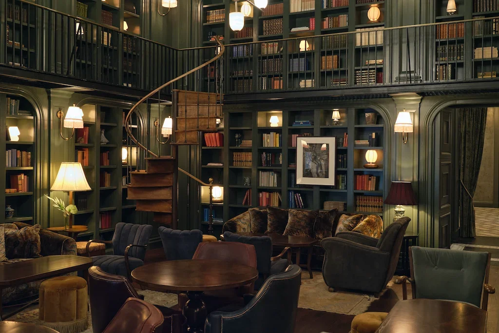 An elegant, well-lit library featuring dark wood bookshelves, a spiral staircase, and comfortable seating with armchairs and sofas.