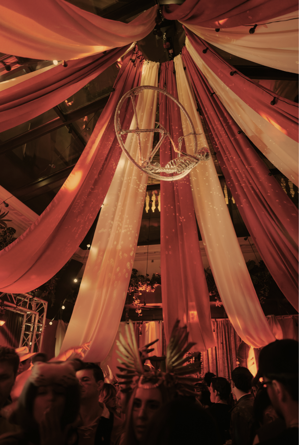 Indoor party scene with pink and white draped fabric ceiling, a hanging chandelier, and a crowd of people.