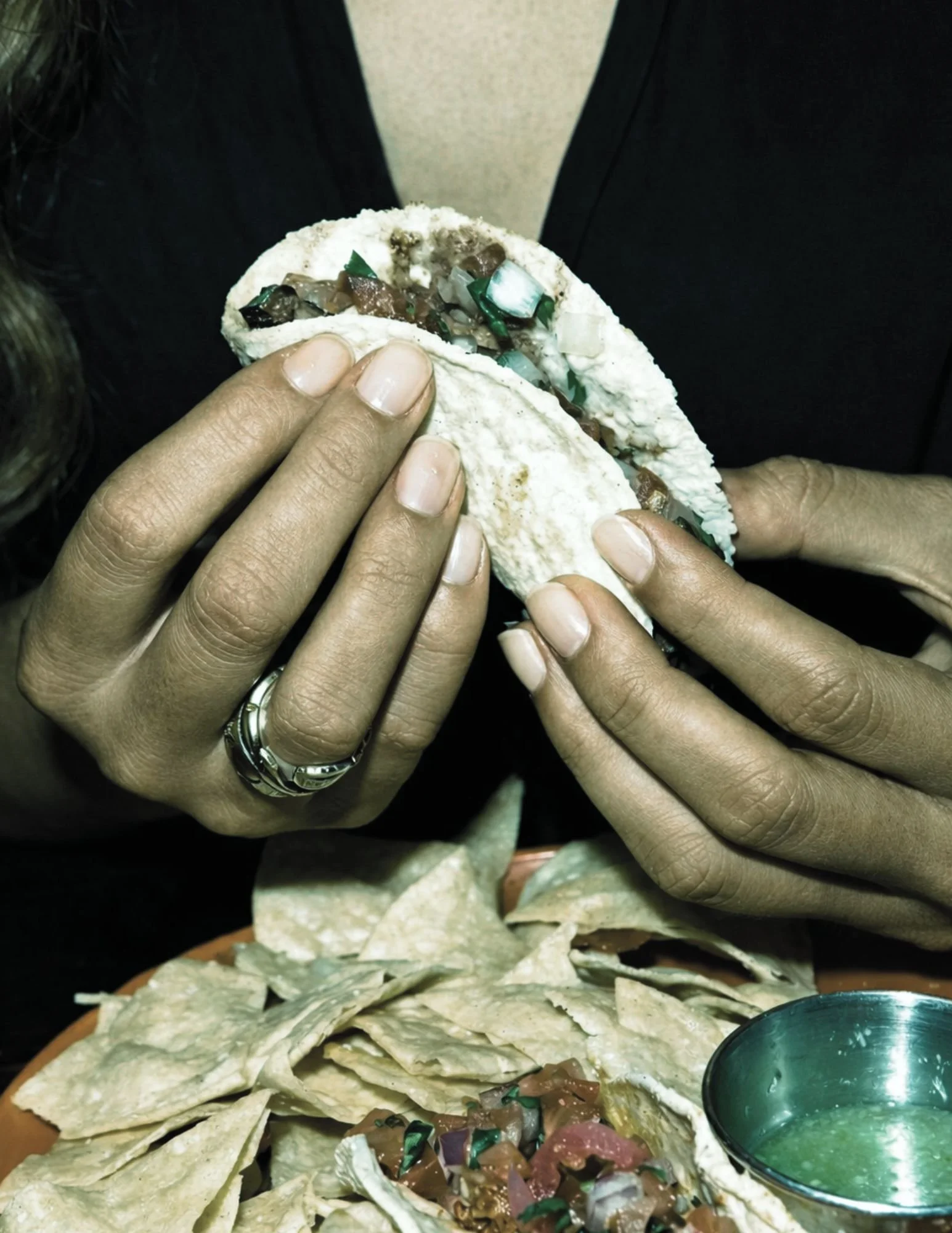 Close-up of hands holding a taco filled with diced onions, green peppers, and beef or meat, over a bed of shredded lettuce with a small metal bowl of green sauce nearby.
