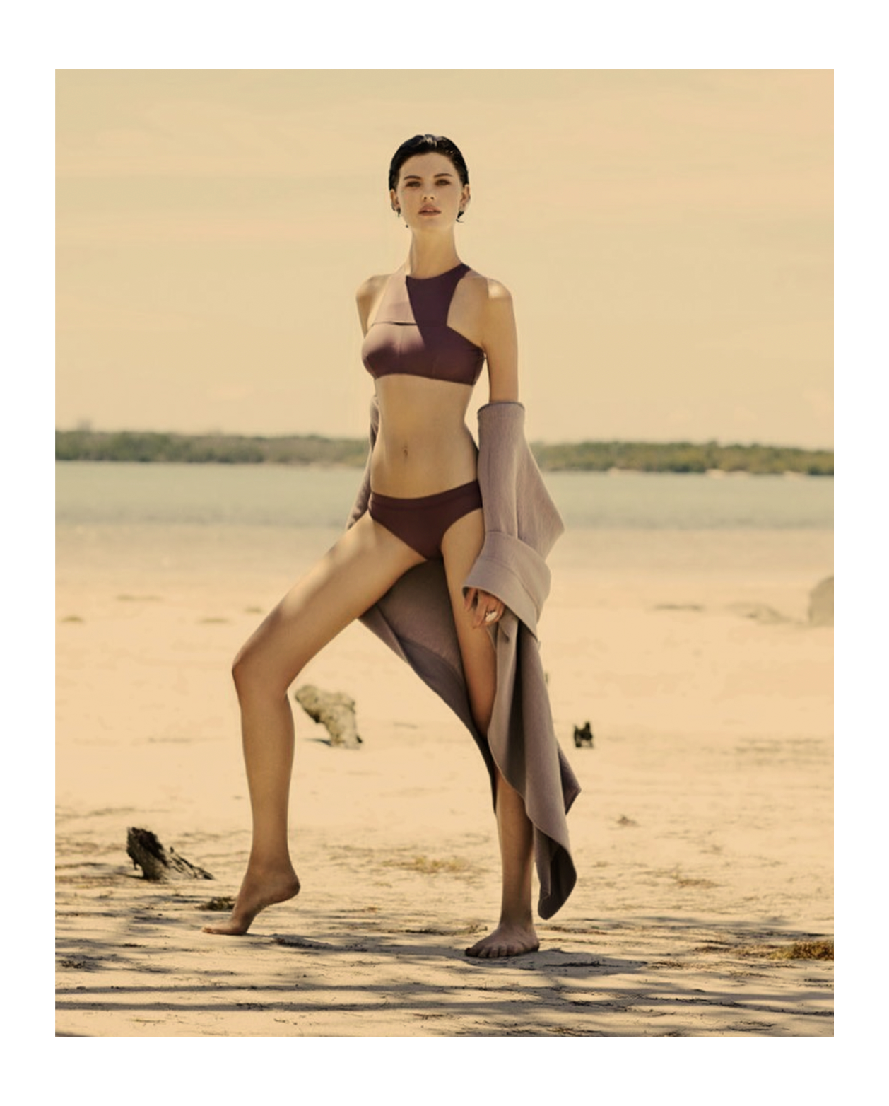 A woman standing on a sandy beach holding a beige coat, wearing a brown bikini with a geometric design, under a clear sky with a distant shoreline in the background.