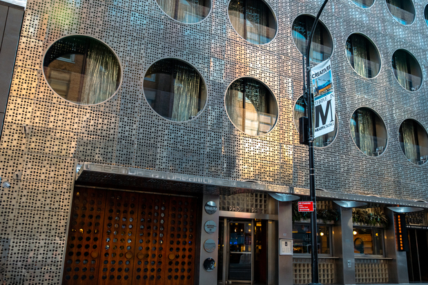 Exterior of a modern building with a perforated metal facade featuring large circular windows, a wooden door, and street signs in front.