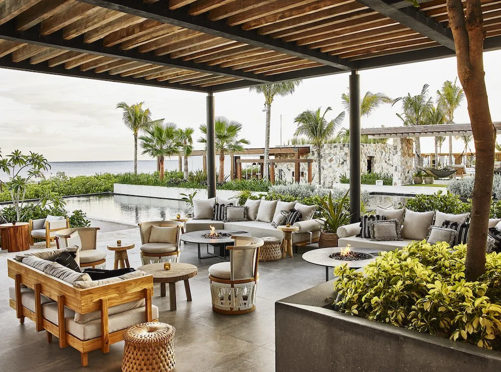 Outdoor seaside lounge area with cushioned seating, coffee tables, and tropical plants under a wooden roof, with palm trees and water in the background.