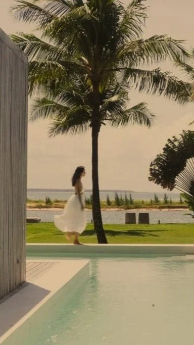 A woman in a white dress walking along a grassy area near a body of water, with a palm tree and a cloudy sky in the background.